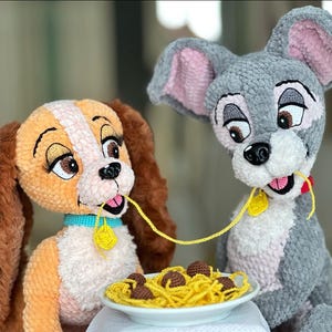 以下が含まれることがあります： 映画「わんわん物語」のレディとトランプの編みぐるみぬいぐるみ。スパゲッティとミートボールの皿を共有しています。レディはタンと茶色、トランプはグレーです。どちらも黄色の首輪タグが付いています。