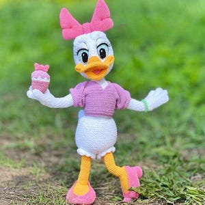 Puede incluir: Mu&ntilde;eca de ganchillo de Daisy Duck hecha a mano, con cuerpo blanco, lazo rosa y top rosa. La mu&ntilde;eca tiene pies amarillos y una magdalena rosa en una mano. La mu&ntilde;eca est&aacute; sobre hierba verde.