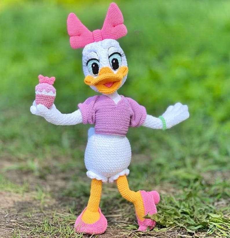 Puede incluir: Mu&ntilde;eca de ganchillo de Daisy Duck hecha a mano, con cuerpo blanco, lazo rosa y top rosa. La mu&ntilde;eca tiene pies amarillos y una magdalena rosa en una mano. La mu&ntilde;eca est&aacute; sobre hierba verde.