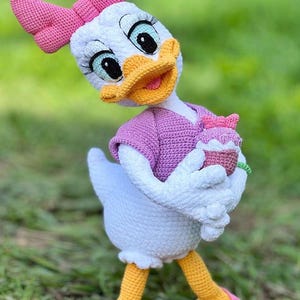 Puede incluir: Mu&ntilde;eca de ganchillo de Daisy Duck hecha a mano, con cuerpo blanco, lazo rosa y top morado. La mu&ntilde;eca sostiene una magdalena y lleva zapatos rosas con suelas amarillas. El fondo es hierba verde borrosa.