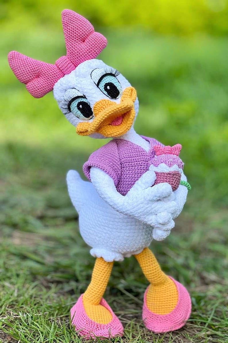 Puede incluir: Mu&ntilde;eca de ganchillo de Daisy Duck hecha a mano, con cuerpo blanco, lazo rosa y top morado. La mu&ntilde;eca sostiene una magdalena y lleva zapatos rosas con suelas amarillas. El fondo es hierba verde borrosa.