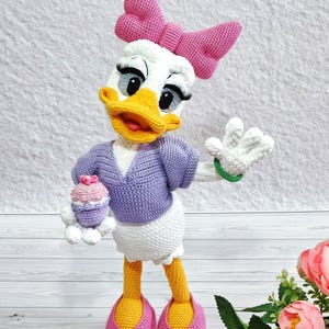 Puede incluir: Mu&ntilde;eca de ganchillo de Daisy Duck, con una cabeza blanca con un lazo rosa, pico amarillo y ojos grandes. Lleva un su&eacute;ter morado, una falda blanca y zapatos rosas. La mu&ntilde;eca sostiene una magdalena y tiene una pulsera verde.
