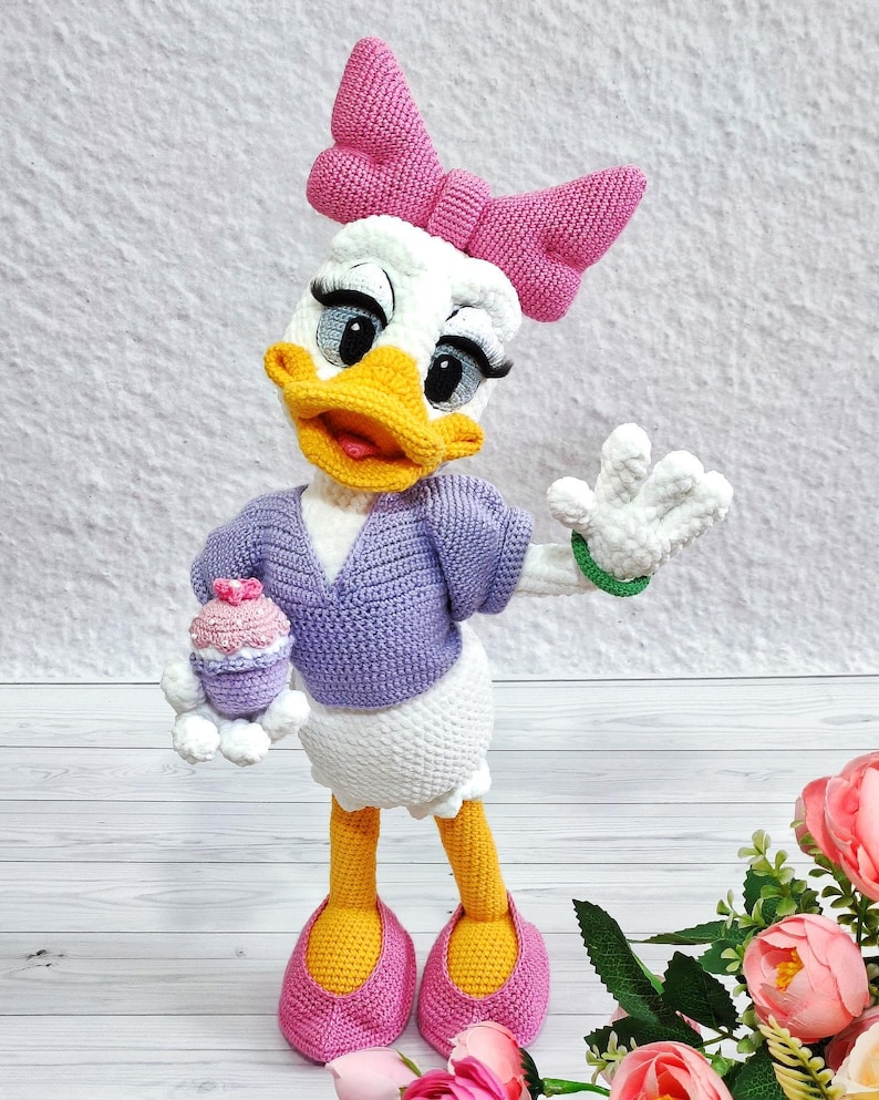 Puede incluir: Mu&ntilde;eca de ganchillo de Daisy Duck, con una cabeza blanca con un lazo rosa, pico amarillo y ojos grandes. Lleva un su&eacute;ter morado, una falda blanca y zapatos rosas. La mu&ntilde;eca sostiene una magdalena y tiene una pulsera verde.