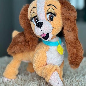 May include: A plush, crocheted dog toy in shades of orange, brown, and white. The dog has large, expressive eyes with long eyelashes, a pink tongue, and a blue collar with a yellow tag. The toy is designed to resemble a cartoon character.
