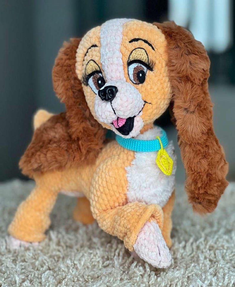 May include: A plush, crocheted dog toy in shades of orange, brown, and white. The dog has large, expressive eyes with long eyelashes, a pink tongue, and a blue collar with a yellow tag. The toy is designed to resemble a cartoon character.