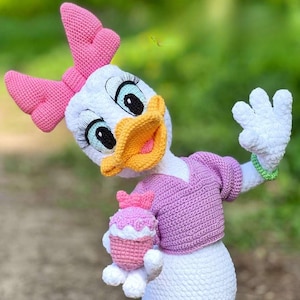 Puede incluir: Una mu&ntilde;eca de ganchillo de Daisy Duck hecha a mano, con cuerpo blanco, lazo rosa y su&eacute;ter rosa. La mu&ntilde;eca tiene pico amarillo, ojos azules y sostiene una magdalena rosa. La mu&ntilde;eca mide aproximadamente 30 cm de altura.