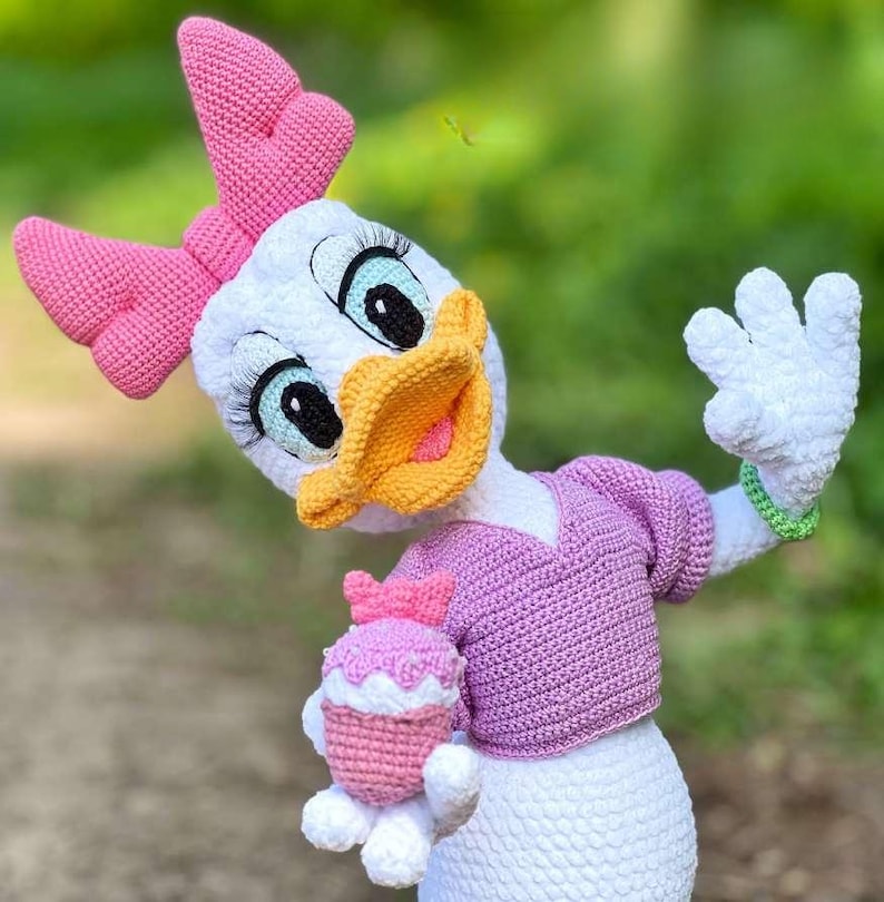 Puede incluir: Una mu&ntilde;eca de ganchillo de Daisy Duck hecha a mano, con cuerpo blanco, lazo rosa y su&eacute;ter rosa. La mu&ntilde;eca tiene pico amarillo, ojos azules y sostiene una magdalena rosa. La mu&ntilde;eca mide aproximadamente 30 cm de altura.
