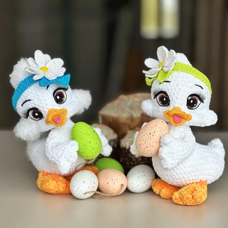 May include: Two white crochet duck figurines with daisy headbands and orange feet. Each duck holds a speckled egg. Easter eggs in various colors and a decorated cake are in the background.