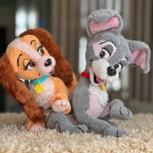 May include: Crocheted plush toys of Lady and Tramp from Lady and the Tramp. Lady is orange and brown with a blue collar and yellow tag. Tramp is gray with pink ears and a red collar. Both are sitting on a cream-colored rug.