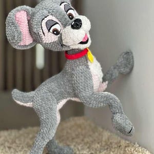 May include: A handmade crochet dog toy in a standing pose. The dog is primarily gray with pink inner ears and a white muzzle. It has brown eyes, a red collar, and a yellow tag. The toy is made of soft yarn and is positioned against a white wall.