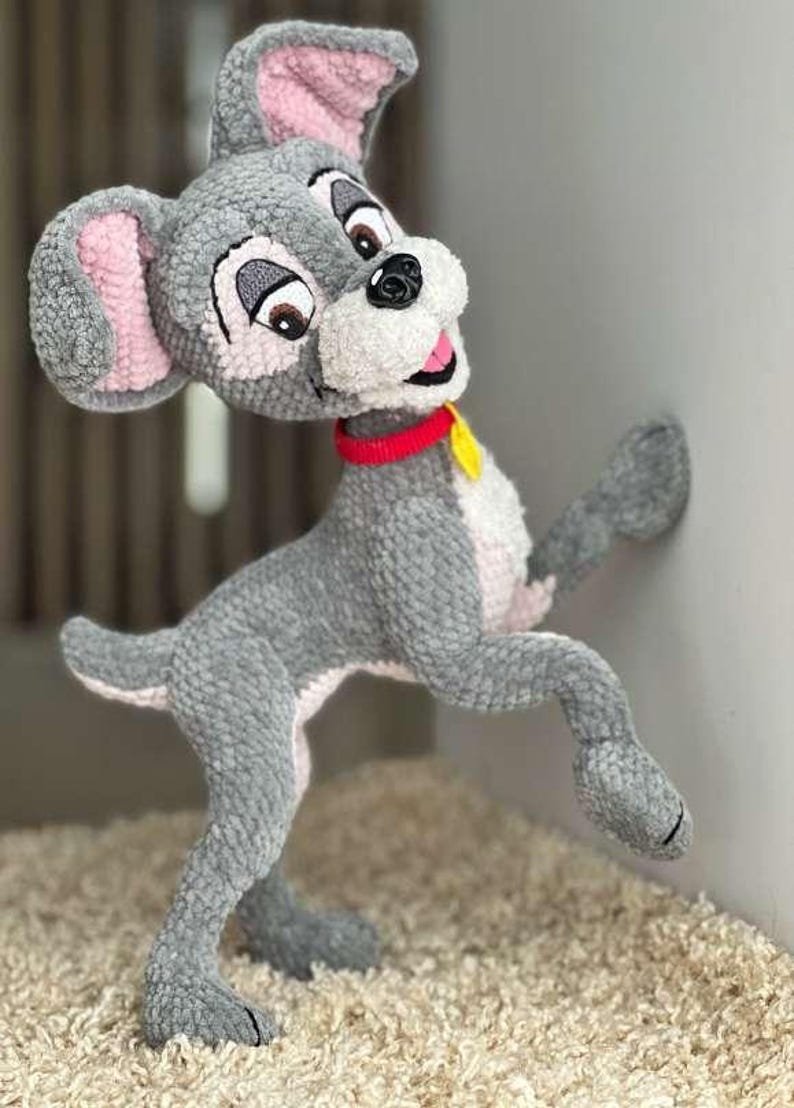 May include: A handmade crochet dog toy in a standing pose. The dog is primarily gray with pink inner ears and a white muzzle. It has brown eyes, a red collar, and a yellow tag. The toy is made of soft yarn and is positioned against a white wall.