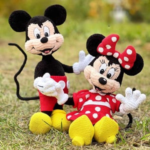 Könnte beinhalten: Zwei gehäkelte Disney-Charakterpuppen, Micky Maus und Minnie Maus, werden auf einer grasbewachsenen Oberfläche gezeigt. Micky trägt rote Shorts, gelbe Schuhe und weiße Handschuhe. Minnie trägt ein rotes Kleid mit weißen Punkten und eine passende Schleife.