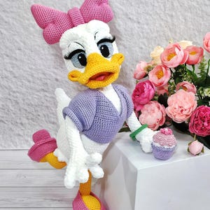 Puede incluir: Mu&ntilde;eca de ganchillo de Daisy Duck hecha a mano con un gran lazo rosa, un su&eacute;ter morado y un peque&ntilde;o cupcake. La mu&ntilde;eca tiene un pico amarillo, pies naranjas y zapatos rosas. Un ramo de flores rosas est&aacute; en el fondo.