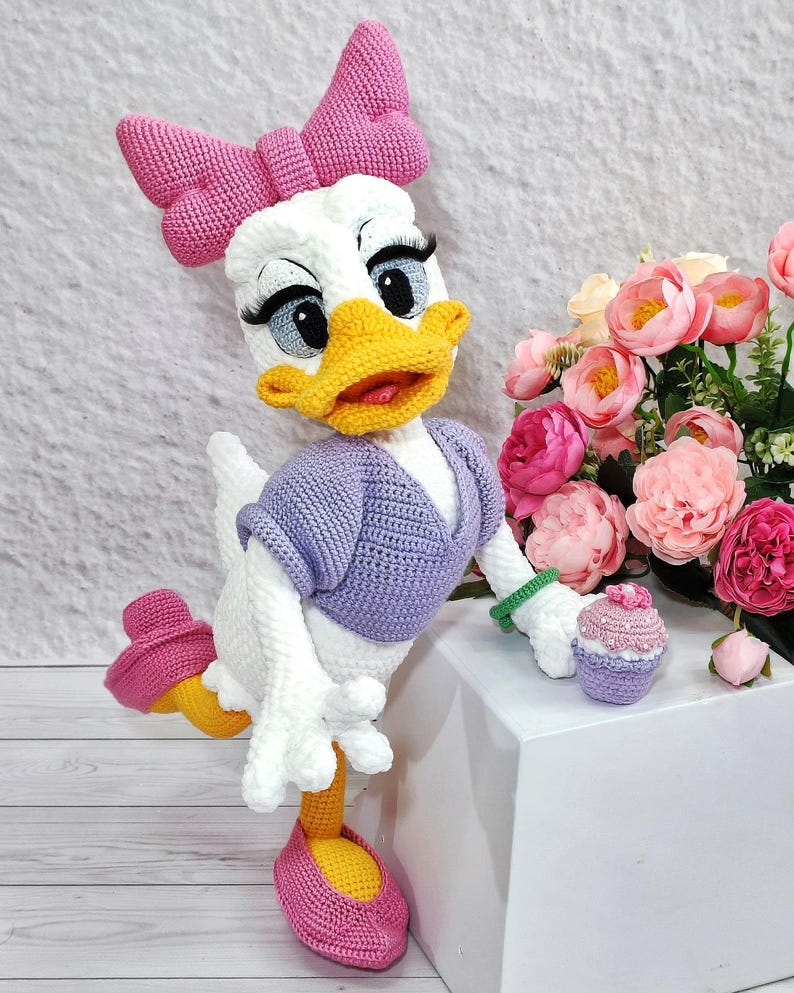 Puede incluir: Mu&ntilde;eca de ganchillo de Daisy Duck hecha a mano con un gran lazo rosa, un su&eacute;ter morado y un peque&ntilde;o cupcake. La mu&ntilde;eca tiene un pico amarillo, pies naranjas y zapatos rosas. Un ramo de flores rosas est&aacute; en el fondo.