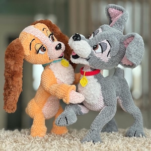 May include: Two plush dog figures, one orange and cream, the other gray, are shown in a playful embrace. The orange dog has long, floppy ears and a blue collar. The gray dog has a red collar. Both have yellow tags. The figures are on a cream-colored surface.