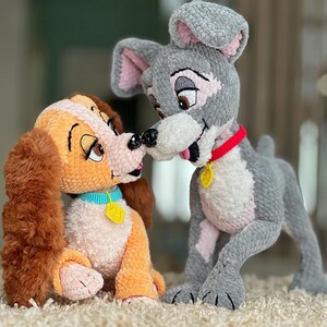 May include: Crocheted Lady and Tramp plush toys. Lady is orange and brown with a blue collar and yellow tag. Tramp is gray with a red collar and yellow tag. Both toys are in a kissing pose, with their noses touching.