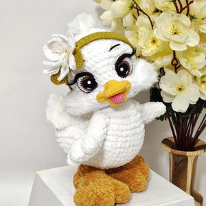 May include: A handmade white crochet duck doll with a yellow beak and feet. The duck has large, expressive eyes with long eyelashes, and a flower and headband. The doll is displayed on a white surface, with yellow flowers in the background.