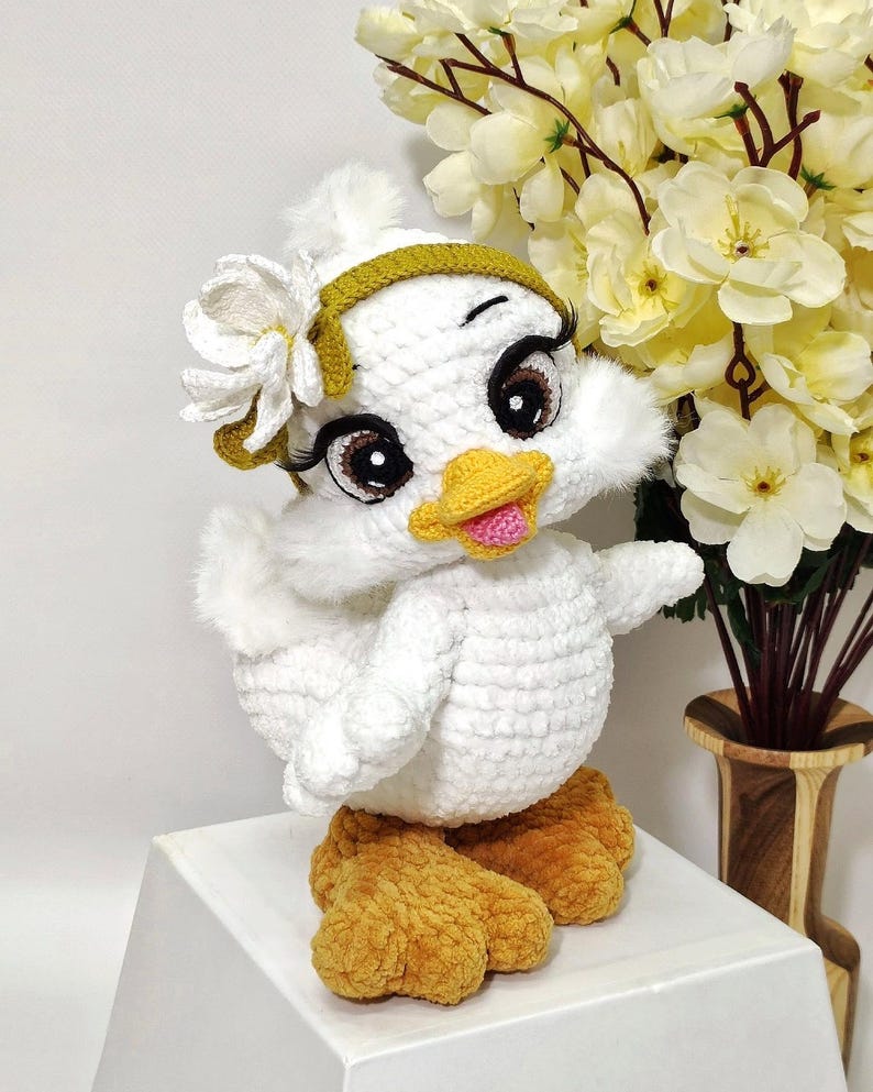 May include: A handmade white crochet duck doll with a yellow beak and feet. The duck has large, expressive eyes with long eyelashes, and a flower and headband. The doll is displayed on a white surface, with yellow flowers in the background.