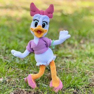 Puede incluir: Mu&ntilde;eca de ganchillo de Daisy Duck hecha a mano. La mu&ntilde;eca tiene un cuerpo blanco, una parte superior morada y un lazo rosa. La mu&ntilde;eca tiene piernas naranjas y zapatos rosas. La mu&ntilde;eca est&aacute; sobre hierba verde.