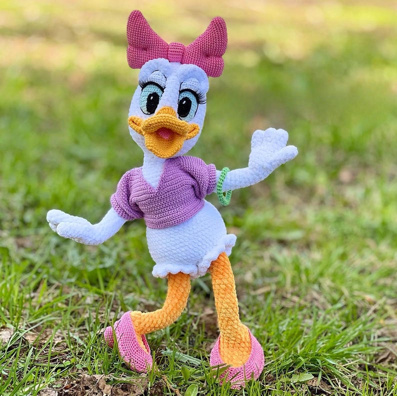 Puede incluir: Mu&ntilde;eca de ganchillo de Daisy Duck hecha a mano. La mu&ntilde;eca tiene un cuerpo blanco, una parte superior morada y un lazo rosa. La mu&ntilde;eca tiene piernas naranjas y zapatos rosas. La mu&ntilde;eca est&aacute; sobre hierba verde.