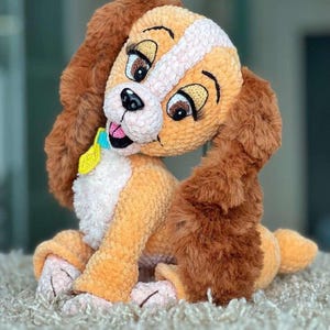 May include: A plush, crocheted dog toy in shades of tan, brown, and white. The dog has large, expressive eyes, long floppy ears, and a small blue and yellow collar. The toy is sitting on a light-colored, textured surface.