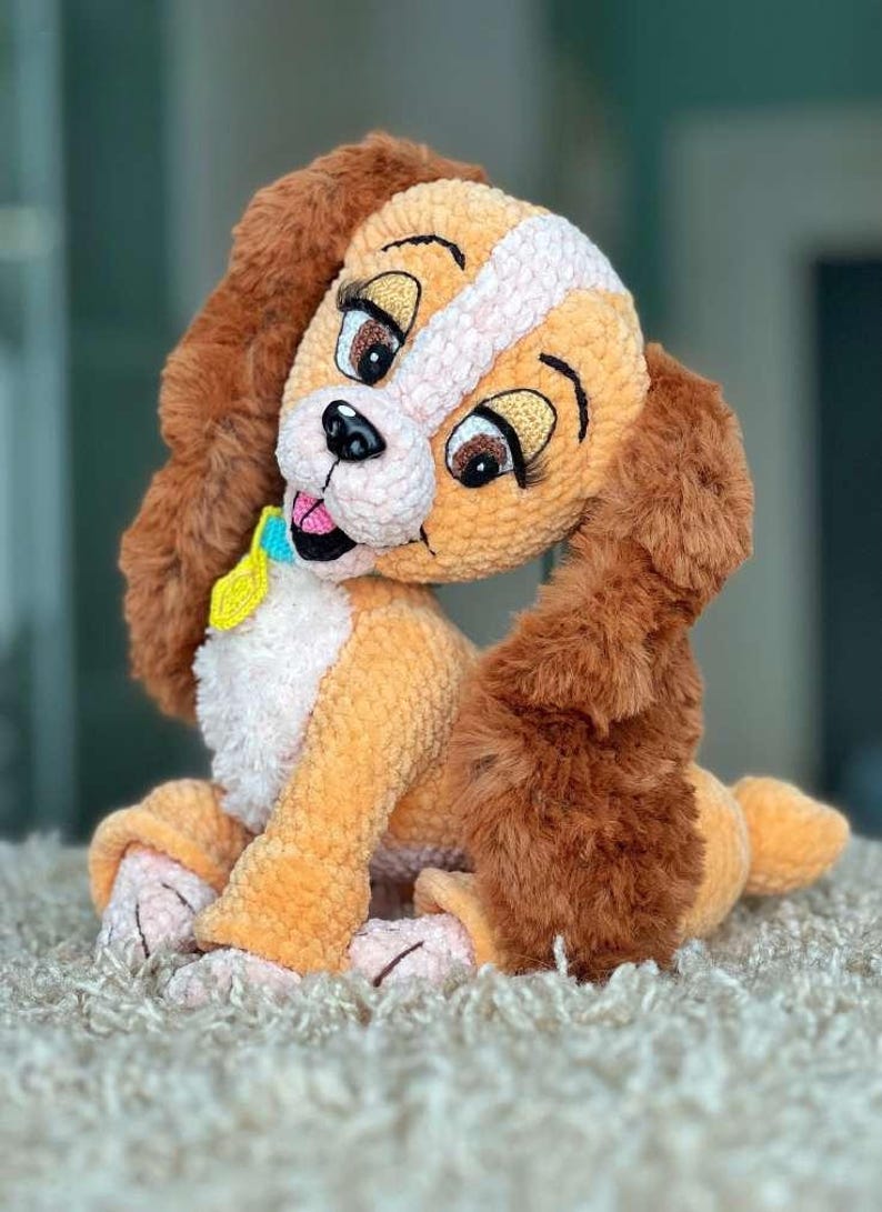 May include: A plush, crocheted dog toy in shades of tan, brown, and white. The dog has large, expressive eyes, long floppy ears, and a small blue and yellow collar. The toy is sitting on a light-colored, textured surface.