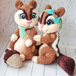May include: Two handmade crochet chipmunk plush toys. The chipmunks are brown, cream, and white with blue eyes and pink noses. One chipmunk wears a green scarf with an acorn, and the other has a blue bow. An acorn is also present.