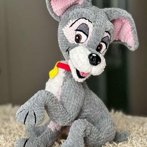 May include: A gray plush dog with pink inner ears, brown eyes, and a red collar with a yellow tag. The handmade stuffed animal is sitting on a beige rug. The dog's features are embroidered.