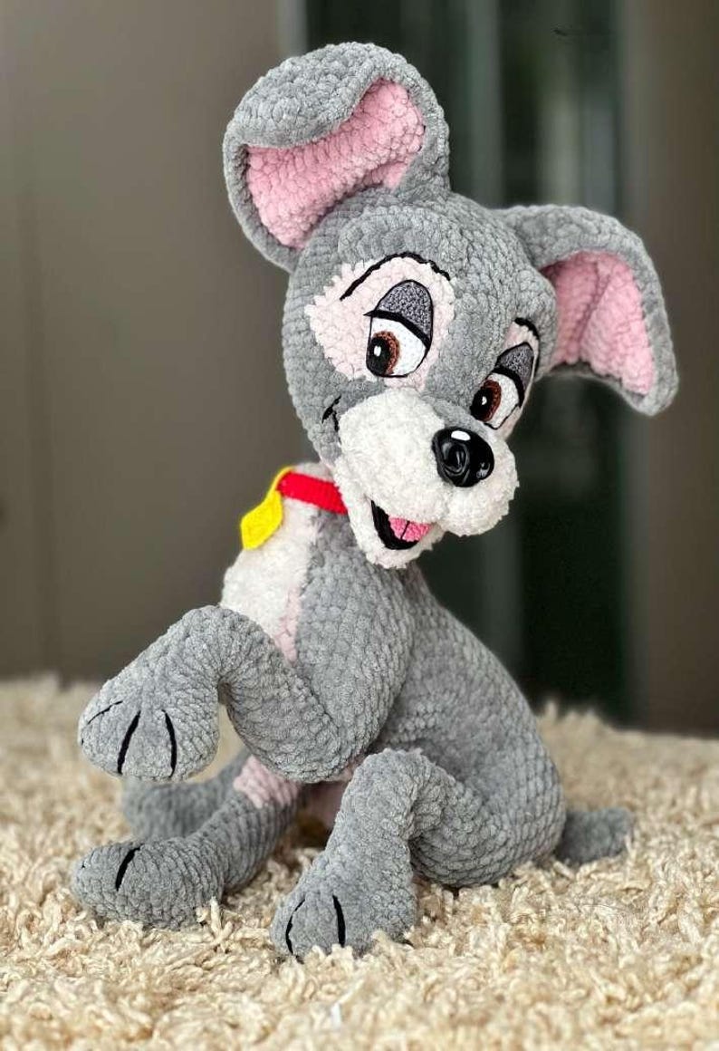 May include: A gray plush dog with pink inner ears, brown eyes, and a red collar with a yellow tag. The handmade stuffed animal is sitting on a beige rug. The dog's features are embroidered.