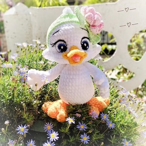 May include: A handmade white crochet duck doll with a green and pink floral hat. The duck has blue eyes, an orange beak, and orange feet. It is sitting in a bed of green plants and small purple flowers.