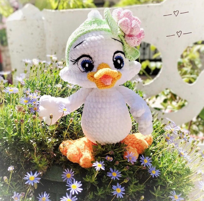 May include: A handmade white crochet duck doll with a green and pink floral hat. The duck has blue eyes, an orange beak, and orange feet. It is sitting in a bed of green plants and small purple flowers.
