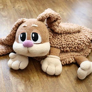 Può includere: Un giocattolo a forma di coniglio di peluche fatto a mano, nei toni del marrone, beige e bianco. Il coniglio ha grandi occhi espressivi, un naso rosa e lunghe orecchie flosce. Il giocattolo è appoggiato su una superficie di legno.