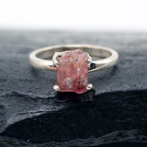 May include: A silver ring featuring a raw, uncut pink gemstone set in a four-prong setting. The stone has a rough, natural texture and a translucent appearance. The band is smooth and metallic, providing a contrast to the irregular shape of the stone.