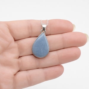 May include: A teardrop-shaped pendant featuring a smooth, matte, dusty blue stone set in a polished silver-toned metal frame. The pendant is attached to a delicate silver-toned chain, creating a simple and elegant piece of jewelry.