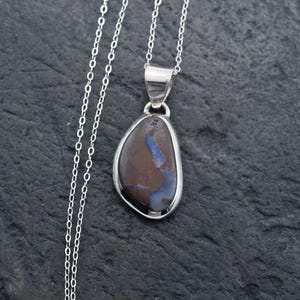 May include: A silver necklace featuring a teardrop-shaped pendant with a dark brown stone inlay that displays a streak of iridescent blue color. The pendant is suspended from a delicate silver chain with a small circular clasp at the end.