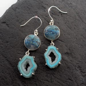 May include: A pair of drop earrings featuring a circular dark blue stone set in silver, suspended above a light blue, irregular geode slice with a crystalline center. The earrings hang from silver ear wires, showcasing a layered design.
