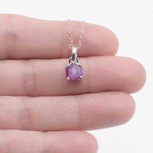 May include: A silver necklace featuring a small, hexagonal-cut, deep pinkish-red gemstone pendant. The stone is set in a simple silver prong setting and hangs from a delicate, thin silver chain, creating a minimalist and refined jewelry piece.