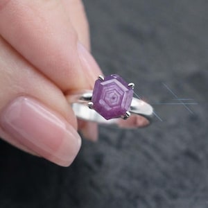 May include: A silver ring featuring a hexagonal, purple-toned gemstone with a distinct geometric pattern on its surface. The stone is held in place by a six-prong setting, and the band is a smooth, polished silver metal, creating a clean look.