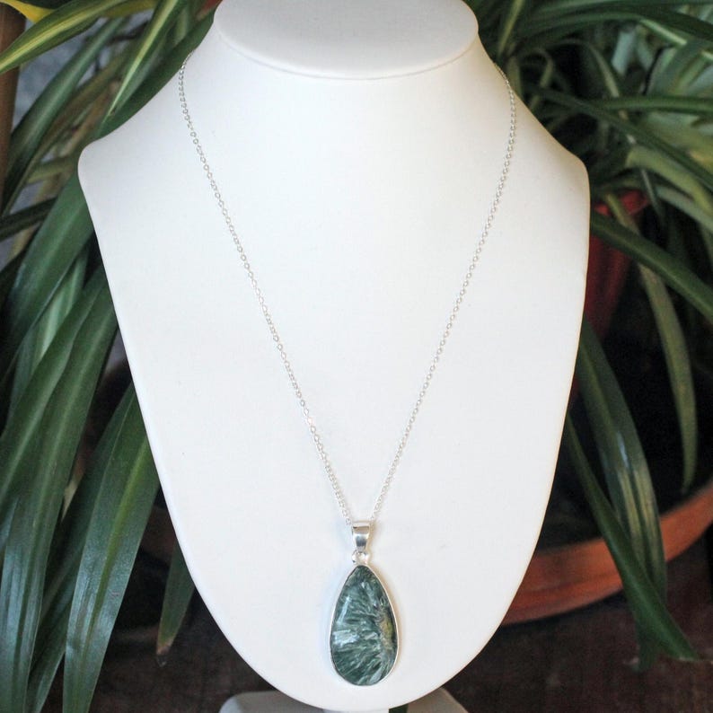 May include: A silver necklace with a teardrop-shaped pendant. The pendant features a green and white stone with a unique, feathery pattern. The necklace is displayed on a white bust against a backdrop of green foliage.