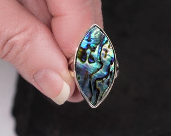 May include: A silver ring featuring a large, marquise-shaped abalone shell. The shell displays iridescent blues, greens, and purples. The ring is held against a dark background, highlighting the vibrant colors of the gemstone.