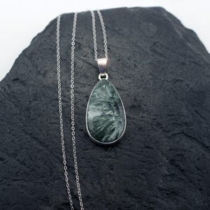May include: A silver necklace featuring a teardrop-shaped pendant. The pendant has a dark green and white marbled stone set in a silver bezel. The chain is a delicate silver link style, and the necklace is displayed on a dark gray surface.