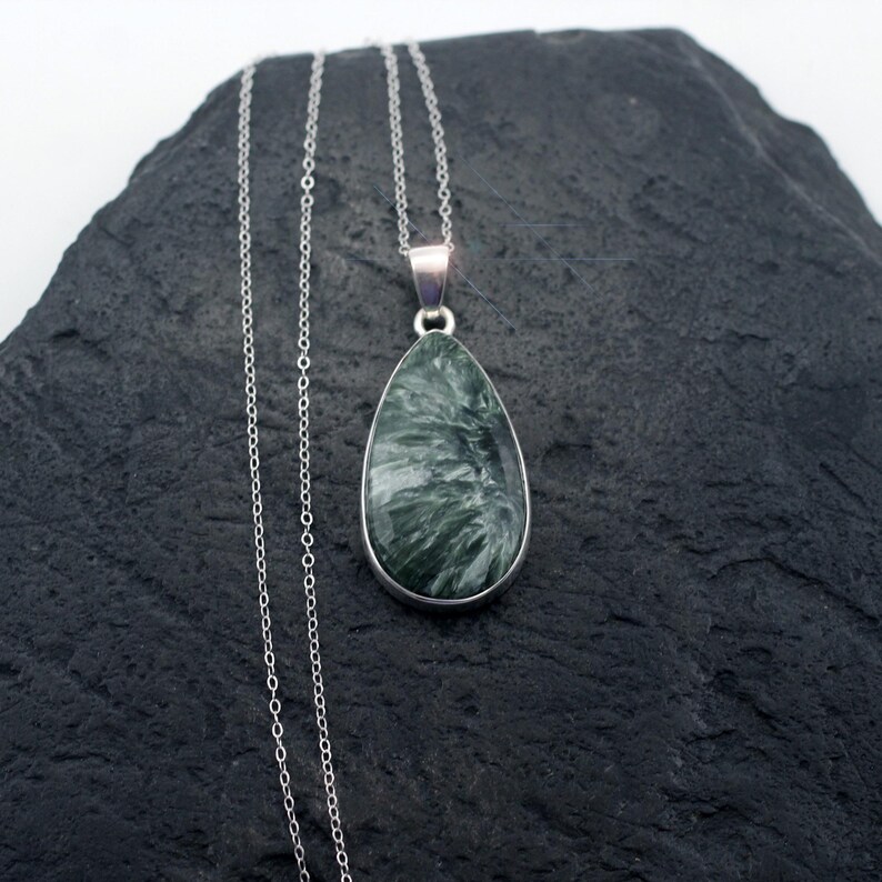 May include: A silver necklace featuring a teardrop-shaped pendant. The pendant has a dark green and white marbled stone set in a silver bezel. The chain is a delicate silver link style, and the necklace is displayed on a dark gray surface.