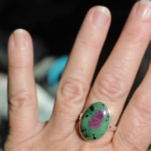 Peut inclure: Bague en argent avec une pierre précieuse ovale. La pierre a un centre rouge, des inclusions vertes et noires. Le bracelet de la bague est en argent poli. La pierre mesure environ 2,5 cm de long.