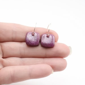 May include: A pair of square-shaped earrings featuring a textured, deep magenta surface with a subtle shimmer. Each earring is accented with a small, clear, prong-set gemstone at the top center, attached to a thin, curved silver-toned wire hook.
