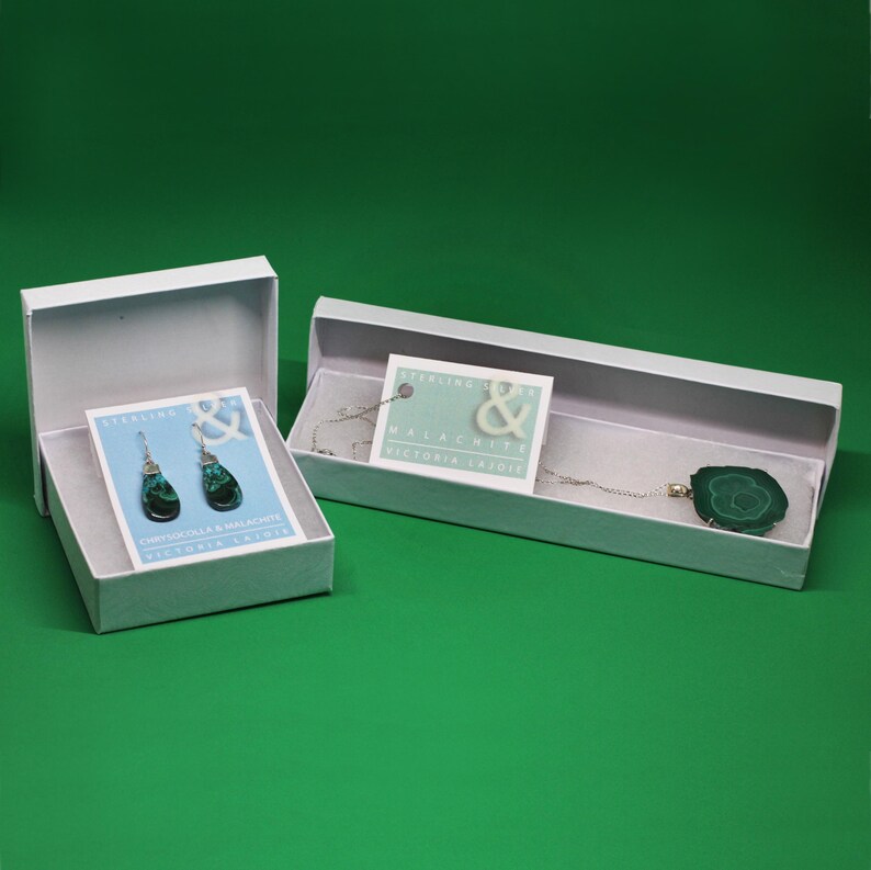 Puede incluir: Dos cajas de regalo blancas con las tapas abiertas. Una caja contiene un par de pendientes de plata de ley con piedras de crisocola y malaquita. La otra caja contiene un collar de plata de ley con un colgante de malaquita. Ambas cajas tienen una peque&ntilde;a tarjeta en el interior con el texto "Sterling Silver & Malachite, Victoria Layder".