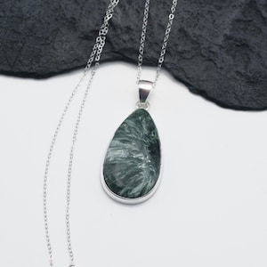 May include: A silver necklace featuring a teardrop-shaped pendant. The pendant has a silver bezel and contains a green and white stone with a unique, swirling pattern. The chain is delicate and hangs on a white surface.