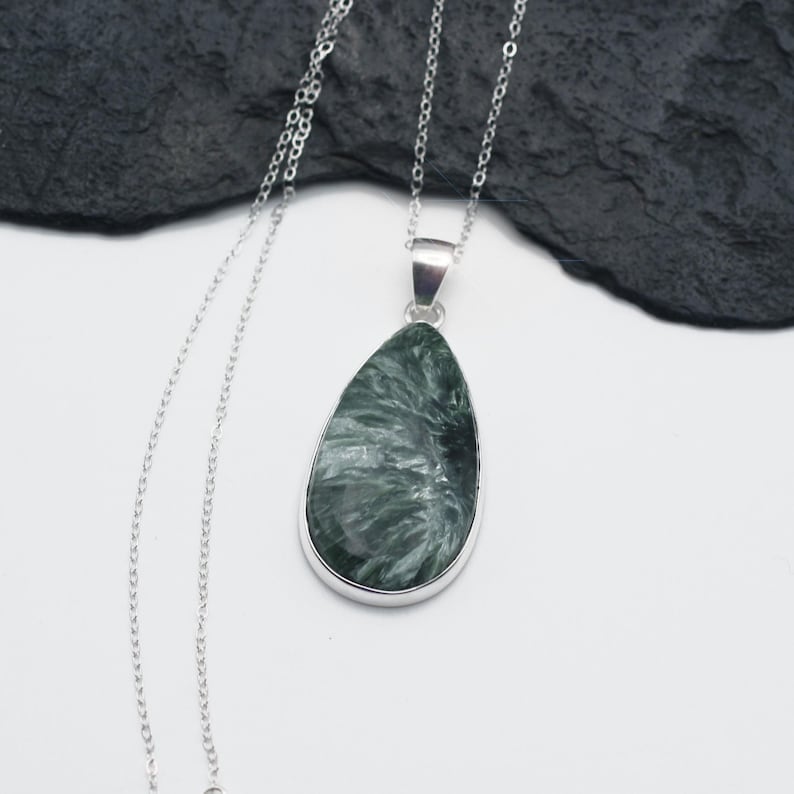 May include: A silver necklace featuring a teardrop-shaped pendant. The pendant has a silver bezel and contains a green and white stone with a unique, swirling pattern. The chain is delicate and hangs on a white surface.