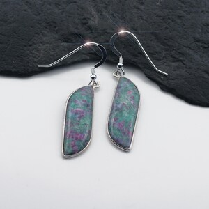 Puede incluir: Un par de pendientes colgantes con una piedra en forma de media luna, enmarcada en metal plateado. La piedra presenta un patrón moteado de tonos verdes y morados, y los pendientes tienen un diseño de gancho simple.