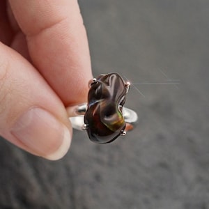 May include: A silver ring featuring a large, irregularly shaped, dark brown gemstone with hints of red and green. The gemstone is held in place by four small prongs. The ring is set against a blurred background.