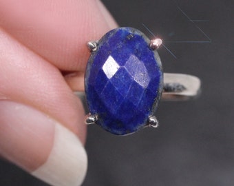 May include: A silver ring featuring a faceted, oval-shaped, deep blue gemstone. The gemstone is held in place by four prongs. The ring is shown against a dark background.